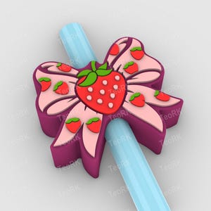 May include: A decorative bow-shaped straw topper featuring a large red strawberry with green leaves and white seeds. The bow is pink and purple with smaller strawberry accents. A light blue straw is inserted through the bow.