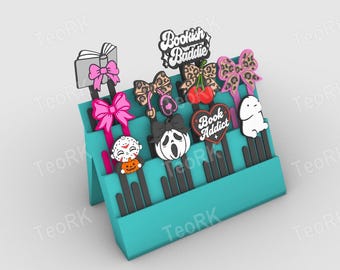 May include: A teal tiered display stand holding several black bookmarks with decorative toppers including a book with a pink bow, leopard print bows, cherries, a heart labeled Book Addict, a ghost, and text reading Bookish Baddie.