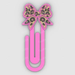 Puede incluir: Un clip rosa con una parte superior en forma de lazo. El lazo presenta un diseño de estampado de leopardo en tonos marrón y beige. El clip es de color rosa brillante.