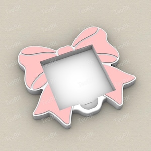 Op de afbeelding: Een roze strikvormig frame met een witte rand en een vierkante opening in het midden. Het frame heeft een 3D-effect, waarbij de details van de strik en de rand duidelijk zijn gedefinieerd. De achtergrond is een neutrale beige kleur.