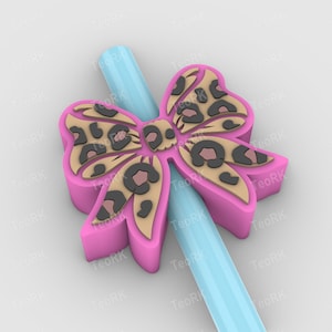 Puede incluir: Un dije en forma de lazo rosa con diseño de estampado de leopardo, colocado sobre una pajita azul claro. El lazo tiene un contorno en relieve y un patrón detallado de manchas negras sobre un fondo beige. El dije es un accesorio decorativo.