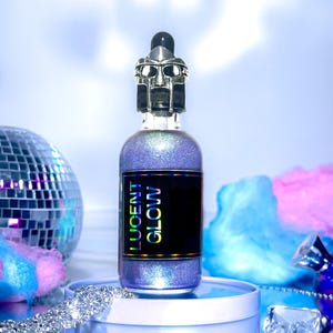 May include: A cosmetic bottle with a silver metal mask-shaped dropper top. The bottle contains shimmering purple liquid and has a black label with "LUCENT GLOW". It sits on a white platform with a mirrored surface. The background includes a disco ball.