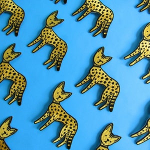 Puede incluir: Una colección de 12 parches bordados de hierro en oro y negro con un diseño de guepardo estilizado. Los parches están dispuestos en un patrón escalonado sobre un fondo azul brillante.