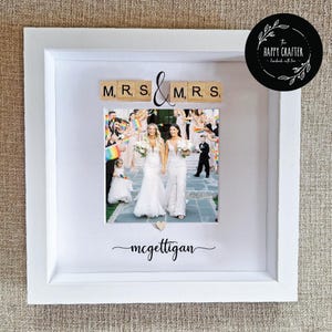 May include: White framed wedding photo featuring two brides. The top of the frame displays "MRS & MRS" in wooden letter tiles. The bottom of the frame has the surname "mcgettigan" written in cursive.