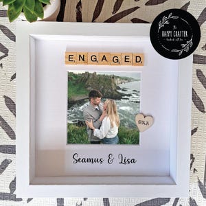 May include: White framed photo featuring a couple's photo with the word "ENGAGED" above. A wooden heart with the date "07.04.24" is included, along with the names "Seamus & Lisa" below the photo, all within the frame.