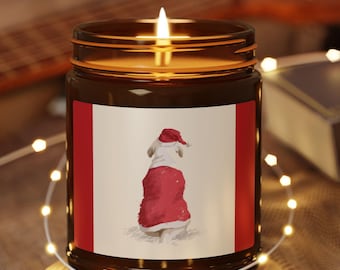Puede incluir: Una vela encendida en un frasco de vidrio marrón con una etiqueta que presenta un perro con un disfraz de Papá Noel. La vela está sobre un posavasos redondo de madera, rodeada de luces de cadena. Una guitarra y un libro están en el fondo.