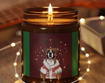 Puede incluir: Una vela encendida en un tarro de cristal marrón con tapa de madera. La etiqueta presenta un perro Boxer con un suéter festivo y cuernos de reno. La vela está sobre un posavasos redondo de madera, rodeada de luces de cadena.
