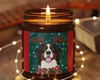 Puede incluir: Una vela encendida en un tarro de cristal marrón con una etiqueta festiva que presenta un perro de montaña bernés con cuernos de reno. La vela está sobre un posavasos de madera, rodeada de luces de hadas.