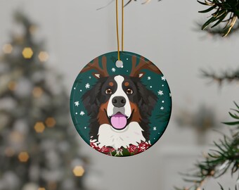 Op de afbeelding: Een ronde kerstversiering met een Berner Sennenhond met rendiergewei en een feestelijke kraag. De hond heeft een zwart, bruin en wit vacht, een roze tong en staat tegen een teal achtergrond met witte sneeuwvlokken.