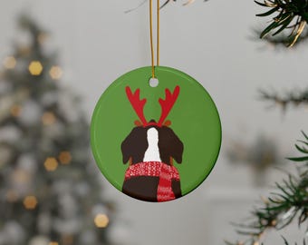 May include: A circular, green Christmas ornament depicting a dog adorned with red reindeer antlers and a red and white scarf. The dog is brown and white. The ornament is suspended by a gold string. A blurred Christmas tree is in the background.