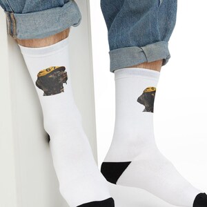 May include: White socks with a black heel and toe, featuring a graphic of a dog wearing a yellow hat. The dog graphic is brown and black. The hat has the number "67" on it. The socks are worn by a person wearing blue jeans.