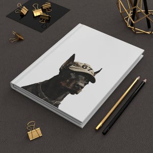 May include: A white journal featuring a Doberman Pinscher wearing a cap with the number 67. The dog is black and brown with a leather collar. Gold and black pencils and gold binder clips are on a dark gray surface.
