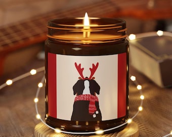 Puede incluir: Una vela encendida en un tarro de vidrio marrón con un diseño festivo. La etiqueta presenta un perro blanco y negro con cuernos de reno y una bufanda roja. La vela está sobre un posavasos de madera, con luces de cadena y una guitarra en el fondo.