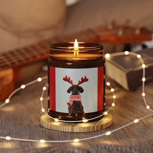 May include: A lit brown glass candle jar featuring a dog wearing reindeer antlers and a red and white sweater. The candle sits on a wooden coaster, with fairy lights around it.