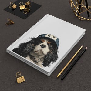 May include: White hardcover journal featuring a Cavalier King Charles Spaniel wearing a denim baseball cap with the number 6.7. The dog has black, brown, and white fur. Gold and black pencils and gold binder clips are on the dark gray surface.