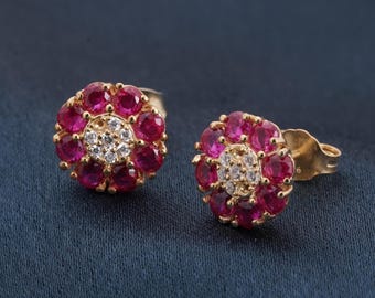 May include: A pair of gold stud earrings. Each earring has a flower design with a ring of red gemstones around a cluster of smaller white gemstones. The earrings are set against a dark blue background.