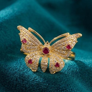 May include: A gold ring shaped like a butterfly, featuring a central round red stone, four smaller round red stones on the wings, and numerous tiny clear pavé-set stones covering the entire surface of the butterfly wings.