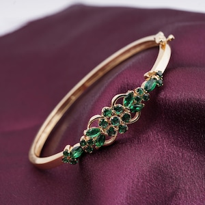 May include: A gold bangle bracelet featuring a floral arrangement of green gemstones. The bracelet is displayed on a deep purple background. The gemstones are different shapes and sizes, and the gold setting provides a contrast.