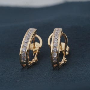 May include: A pair of gold-colored hoop earrings. Each earring features a row of clear, sparkling stones set along the outer curve. The earrings have a classic, elegant design.