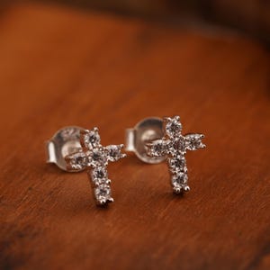 Può includere: Un paio di orecchini a bottone color argento, ciascuno a forma di croce e incastonato con piccole pietre preziose trasparenti. Gli orecchini sono esposti su una superficie di legno marrone caldo.