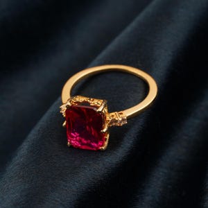 May include: A gold ring featuring a large, rectangular, deep red gemstone. The ring has an ornate design around the gemstone and small accent stones on the band. The ring is set against a dark background.