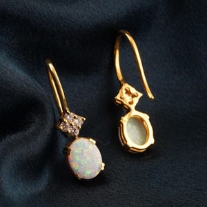 May include: A pair of gold-colored drop earrings. Each earring features an oval, iridescent opal stone set in a gold frame. The top of each earring has a small, diamond-studded accent. The earrings hang from a curved gold hook.