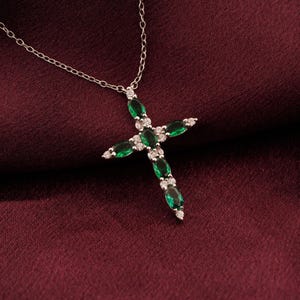 May include: A silver-toned cross pendant necklace with a delicate chain. The cross is adorned with oval-cut emerald green gemstones and small, sparkling white stones. The necklace rests on a deep burgundy fabric.