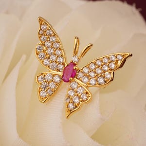 May include: A gold-colored butterfly brooch with clear, round gemstones and a central oval-shaped ruby. The butterfly's wings are detailed with multiple small gemstones, and the body features a ruby.