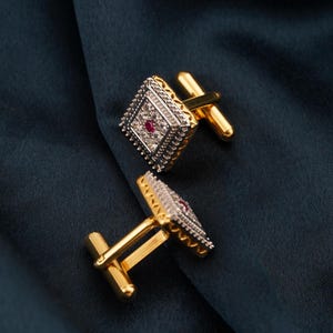 May include: A pair of gold and silver cufflinks with a diamond-shaped design. Each cufflink features a central red gemstone surrounded by smaller white stones and a decorative border. The cufflinks have a gold bar for attachment.