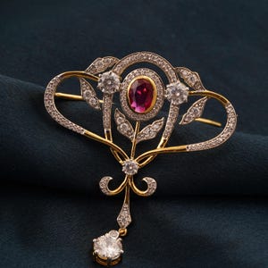 Peut inclure: Broche en or et argent avec un rubis ovale central et des pierres précieuses claires. La broche présente un motif floral avec une pierre précieuse pendante en bas. Le fond est de couleur teal foncé.