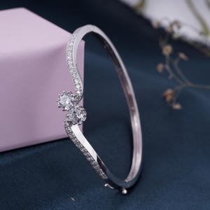 Op de afbeelding: Een zilverkleurige armband met een gebogen ontwerp. De armband heeft een reeks kleine, heldere stenen langs de gebogen band. Twee grotere, ronde, heldere stenen zijn in het midden van het ontwerp geplaatst en vormen een blikvanger.