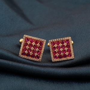 May include: Two square gold cufflinks, each featuring a pattern of red and white gemstones. The cufflinks have a gold border and are set against a dark teal background.