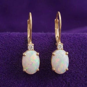 May include: A pair of gold-colored leverback earrings. Each earring features an oval, iridescent opal stone and a small, clear gemstone accent. The earrings are displayed against a purple background.