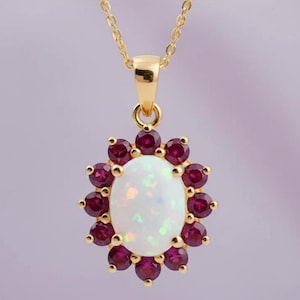 Könnte beinhalten: Goldfarbene Halskette mit Anhänger. Der Anhänger hat einen ovalen, weißen Opal, umgeben von kleinen, runden, dunkelroten Edelsteinen. Die Kette hat eine feine Kette und einen einfachen Verschluss.