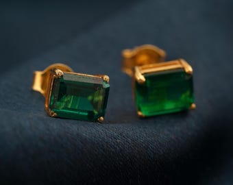Pode incluir: Um par de brincos de botão dourados, cada um com uma pedra preciosa retangular verde esmeralda. Os brincos são apresentados sobre um fundo azul escuro, realçando o verde vibrante das pedras.
