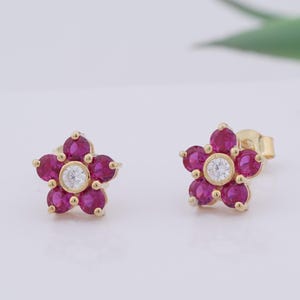 May include: A pair of gold flower stud earrings. Each earring features five magenta-colored gemstones arranged around a central white gemstone. The earrings are set on a light background.