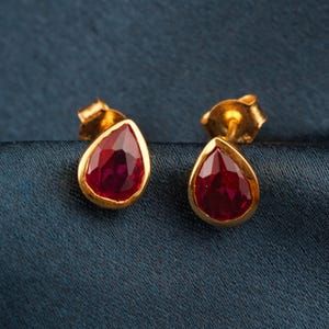 May include: A pair of gold stud earrings with teardrop-shaped, faceted, deep red gemstones. The gemstones are set in a gold bezel, and the earrings are displayed on a dark blue fabric background.