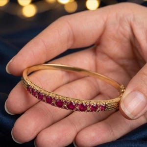 Może przedstawiać: Złota bransoletka typu bangle z zawiasem, ozdobiona rzędem okrągłych, ciemnoczerwonych kamieni szlachetnych z przodu. Metalowa obręcz posiada misternie grawerowane detale po bokach, nadające polerowanej złotej powierzchni teksturę.