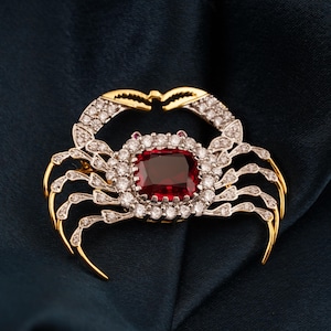 May include: A crab-shaped brooch with a large, square, red gemstone at its centre. The brooch features gold-coloured claws and legs, and the body is encrusted with small, sparkling white gemstones. The crab is set against a dark blue background.