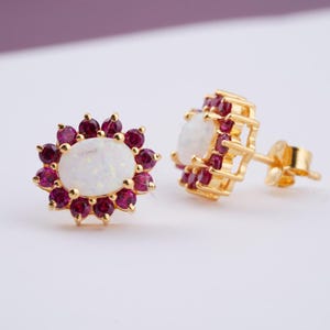 May include: A pair of gold-coloured stud earrings. Each earring features a central oval-shaped white gemstone surrounded by small, round, deep red gemstones. The earrings have a floral design and are set against a soft, neutral background.