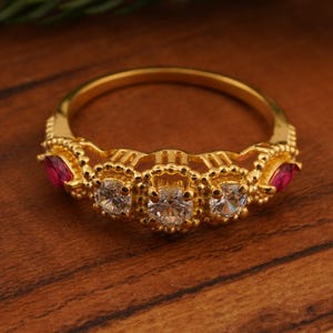 May include: A gold-colored ring featuring three clear, round gemstones in the center, flanked by two red gemstones. The ring has a detailed, ornate design with a textured band. The ring is displayed on a wooden surface.