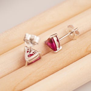 May include: A pair of silver stud earrings with triangular, deep pink gemstones. The earrings are set in a silver frame and have a post and butterfly backing. The earrings are displayed on a light brown wooden surface.