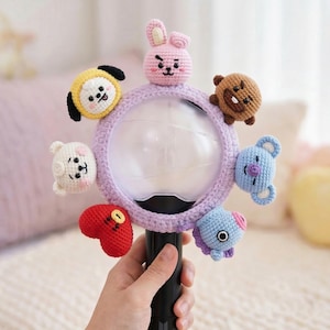 May include: A black and purple light stick with a crocheted lavender ring featuring seven colourful, cartoon-like characters. The characters are in various colours, including yellow, pink, brown, blue, white, and red. The light stick is held by a person.