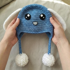 May include: A handmade blue crochet hat with white accents, featuring a cartoon face with black eyes and a smiling mouth. The hat has white bunny ears and dangling blue braids with white pom-poms. A whimsical and cozy accessory.