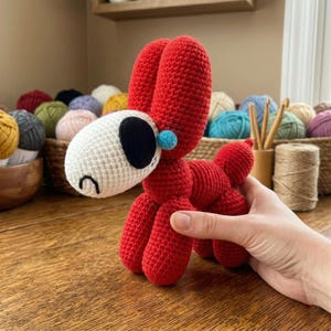 May include: A handmade red crochet dog with a white head, black eye, and blue accents. The dog is made of yarn and has a whimsical design. The background includes yarn balls and crochet supplies.