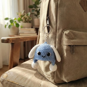 May include: A blue crocheted hat keychain with white ear-like accents and two white pom-poms. The keychain is attached to a silver keyring and hangs from a tan backpack. The hat has a smiling face with black eyes.