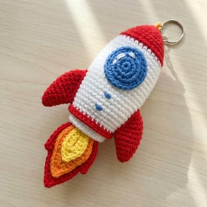 May include: A handmade crochet rocket ship keychain in red, white, blue, orange, and yellow. The rocket has red wings and a flame-like tail. The rocket's body is white with a blue circle and blue dots. A silver keyring is attached.