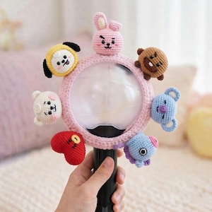 May include: A pink and white crochet light stick with seven small, colorful crochet characters attached around the light. The characters include a yellow circle, a white bear, a red heart, a blue creature, a brown bear, a blue bear, and a pink bunny.
