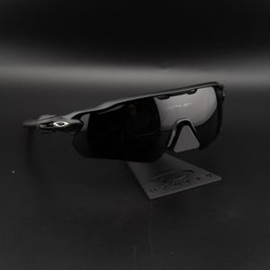 May include: Black Oakley sunglasses with a sleek, modern design. The sunglasses have a glossy finish and are displayed on a black stand. The Oakley logo is visible on the frame.