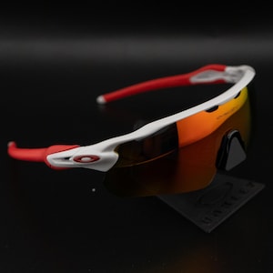 May include: White and red sports sunglasses with orange-tinted lenses. The sunglasses have a sleek, modern design with a white frame and red accents on the arms. The lenses are reflective and appear to be made of a durable material.